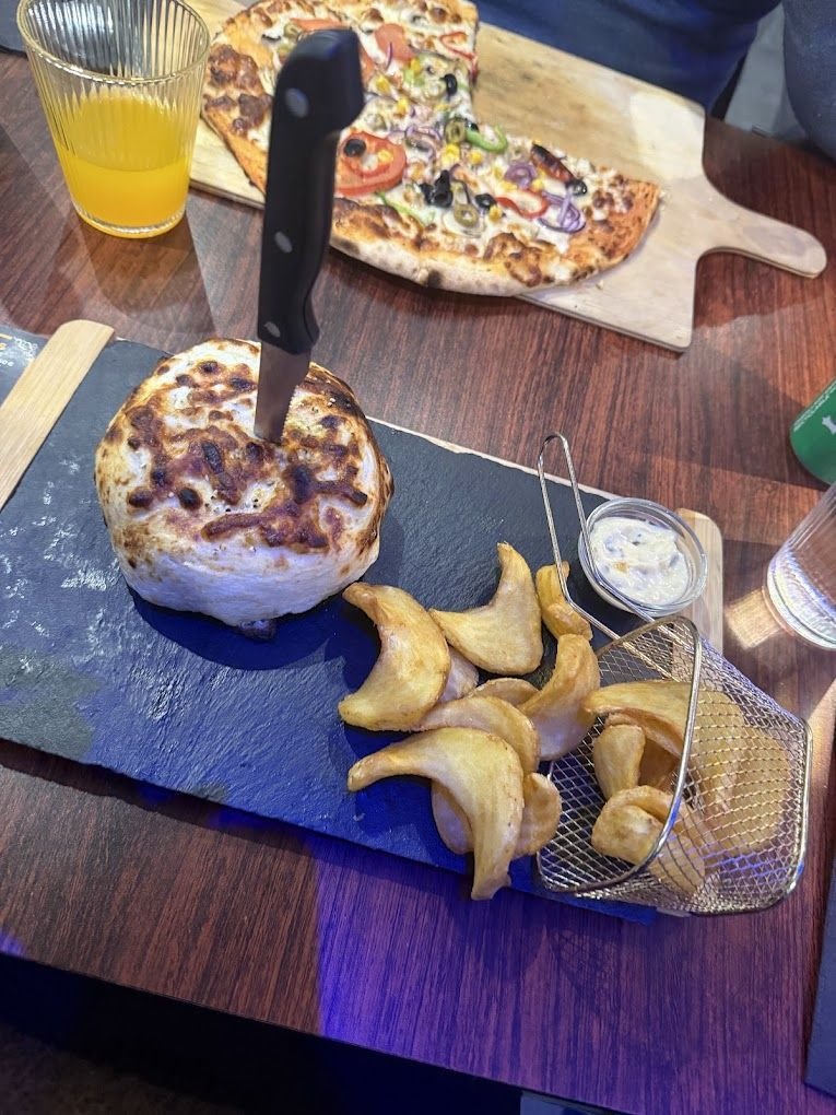 Spécialité gratinée au four servie sur ardoise avec potatoes, pizza en arrière-plan
